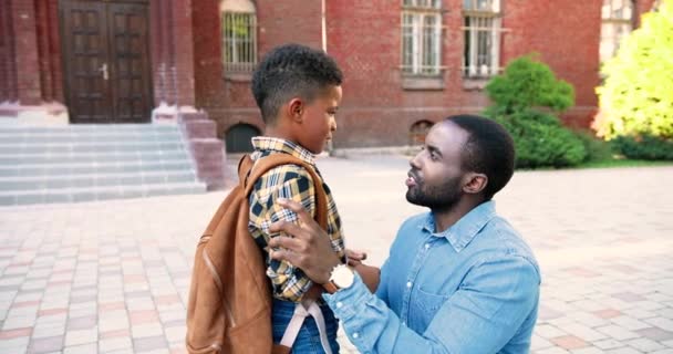 Πορτρέτο του όμορφου Αφροαμερικανού νεαρού που μιλάει με τον χαριτωμένο γιο του στην αυλή του σχολείου έξω. Καλός μαθητής με σχολική τσάντα που μιλάει με τον πατέρα κοντά στο σχολείο. Οικογενειακή έννοια — Αρχείο Βίντεο