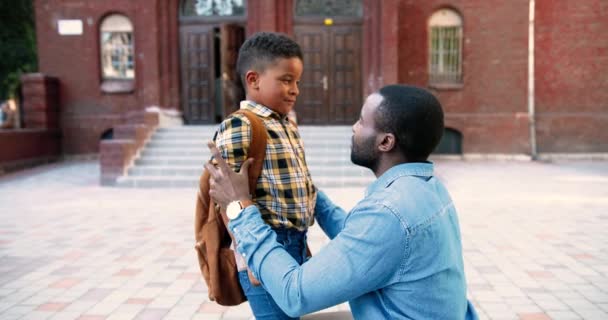 Retrato de homem afro-americano bonito falando e animando o filho no pátio da escola ao ar livre. Feliz adolescente estudante júnior com saco escolar falando com o pai perto da escola. Conceito familiar — Vídeo de Stock