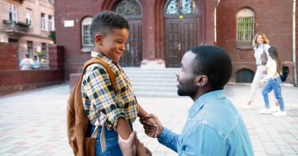 Retrato de homem afro-americano bonito falando e animando o filho no pátio da escola ao ar livre. Aluno macho feliz com saco escolar falando com o pai perto da escola. Menina caucasiana com a mãe no fundo — Vídeo de Stock