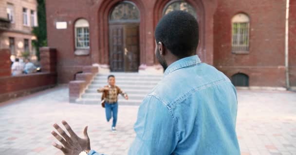Портрет красивого афроамериканца, встречающего сына из школы и обнимающего его на школьном дворе. Счастливый ученик мужского пола разговаривает с отцом возле школы. Кавказская девушка бежит на заднем плане — стоковое видео