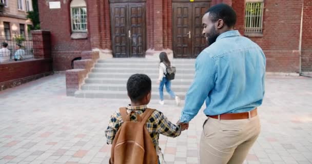 Pais multi-étnicos trazem crianças para a escola. Uma estudante caucasiana a correr para a escola. Pai afro-americano conversando com menino perto da escola de bom humor. O estudante corre para as aulas. Conceito do estudo — Vídeo de Stock