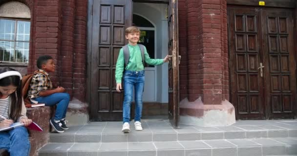 Joyful Caucasian boy leaving school and meeting mother outdoors. Beautiful woman hugging schoolboy. African American kid sitting and pretty girl writing into planner near school. Pupils concept — Stock Video