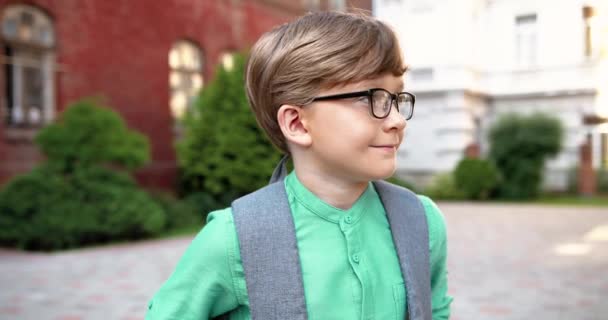 Gros plan portrait de l'écolier mignon heureux dans des lunettes détournant les yeux et souriant à la caméra. Joyeux adolescent caucasien de bonne humeur à la cour d'école. Des émotions positives. Étudiant junior. Concept de sourire — Video