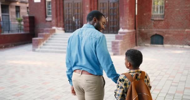 Posteriore di bell'uomo afroamericano che cammina con uno scolaro carino vicino a scuola e chatta all'aperto. Allievo adolescente maschio con lo zaino che parla con suo padre. Relazioni padre-figlio. Concetto familiare — Video Stock