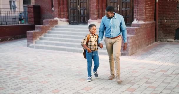 幸せなハンサムなアフリカ系アメリカ人の男が学校の近くにかわいい小学生と歩いて話しています。クラスの後に彼のお父さんと屋外でチャットバックパックを持つ10代の中学生。会話の概念 — ストック動画