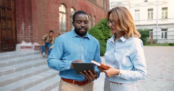 Portrait of joyful mixed-races teachers talking and tapping on tablet outdoors. Handsome African American man and Caucasian pretty woman chatting and typing on device at schoolyard. School concept — Stock Video