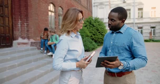Portrait of joyful mixed-races teachers talking outdoor. Handsome African American man tapping on tablet and speaking with Caucasian pretty woman educator at schoolyard with pupils on background. — Stock Video