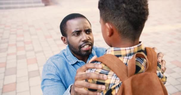 学校の庭で少年と話している深刻な不幸なアフリカ系アメリカ人男性の肖像画を閉じます。悪い気分で父は彼の10代の息子の屋外と口論する。バックパック付きの学童。会話の概念 — ストック動画