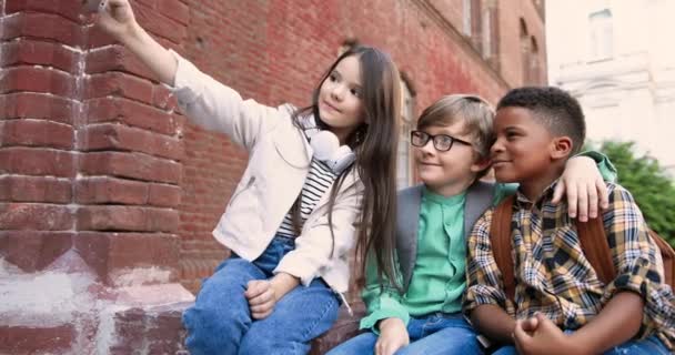 Niños felices de razas mixtas sentados cerca de la escuela y tomando fotos en el teléfono inteligente al aire libre. Caucásico chica bonita haciendo foto selfie con niño en gafas y compañero de clase afroamericano en el teléfono celular. — Vídeos de Stock