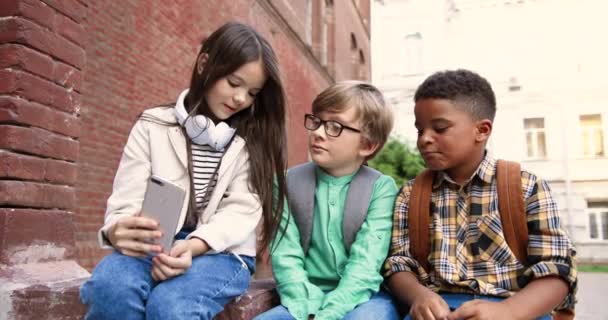 Porträtt av blandade raser barn sitter nära skolan och pratar utomhus. Vit söt flicka håller smartphone i handen och chatta med pojke i glasögon och afroamerikansk klasskamrat. Vänner — Stockvideo