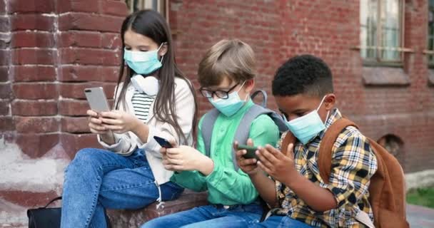 Porträtt av blandraser barn i masker sitter nära skolan och knacka på mobiltelefoner utomhus. Vit söt flicka som använder smartphone. En afroamerikansk skolpojke som surfar på smartphone. Vänner — Stockvideo