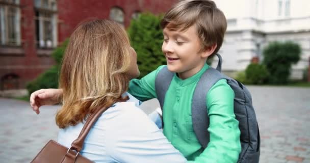 Zamknij portret pięknej białej kobiety przytulającej syna na podwórku szkolnym. Wesoły nastolatek mężczyzna junior student z torbą szkolną przytula matkę w pobliżu szkoły. Pojęcie miłości — Wideo stockowe