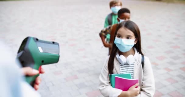 Dekat dengan tangan wanita Kaukasia memegang termometer elektronik dan mengukur suhu murid sebelum kelas. Gadis cantik dengan notebook. Anak sekolah Afrika-Amerika bertopeng. Konsep Virus — Stok Video