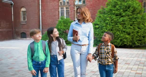 Szczęśliwa biała piękna nauczycielka stojąca z uczniami na zewnątrz i uśmiechnięta. Kobieta pedagog z planistą w ręku rozmawia z dziewczynami i chłopcami mieszane wyścigi młodszych studentów. Koncepcja edukacji — Wideo stockowe