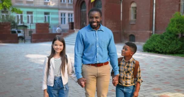 Glada stilig afroamerikansk manlig pedagog som står med multietniska barn nära skolan och ler. Glad man lärare på gott humör med söt vit flicka och liten skolpojke Studie koncept — Stockvideo