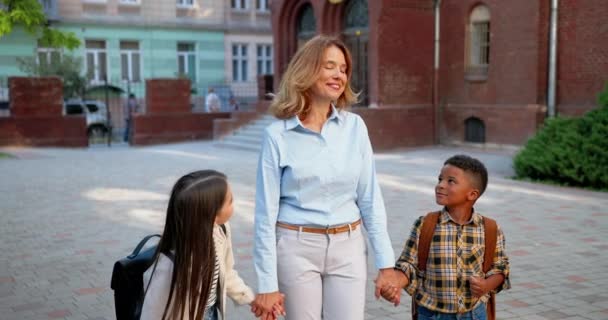 Joyful Caucasian beautiful female teacher standing with pupils at schoolyard and smiling. Woman educator looking at girl and African American boy outdoor. Mixed-races junior students. School concept — Stock Video