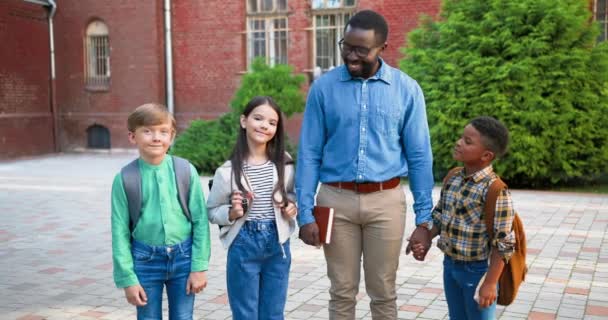 Gelukkige Afro-Amerikaanse mannelijke leraar met multi-etnische leerlingen buiten en glimlachend. Man opvoeder met blank meisje en jongen buiten. Vrolijke studenten. Onderwijsconcept — Stockvideo