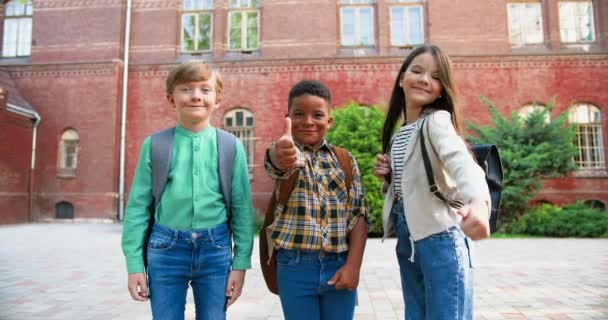 Vrolijke gemengde-rassen junior studenten met schooltassen staan voor de school en het geven van duimen omhoog. Mooi blank meisje glimlachend buiten. Afro-Amerikaanse knappe jongen in goed humeur. Leerlingconcept — Stockvideo