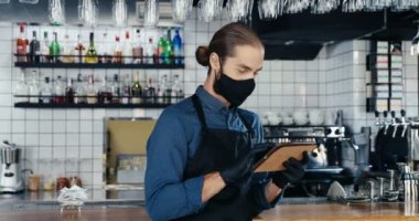 Maskeli, eldivenli ve önlüklü beyaz bir erkek mutfaktaki kafede tablet kullanıyor. Bar 'daki yakışıklı garson tablet bilgisayarda internete bağlanıp kayıyor. Gadget kullanıcısı. Karantina.