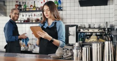 Önlüklü, kafeteryada duran, tablet cihazı kullanan ve erkek garsonla konuşan güzel, beyaz bir bayan garson. Kadın barikat tıklatıyor ve barmenle sohbet ederken zımbırtıyı kaydırıyor..