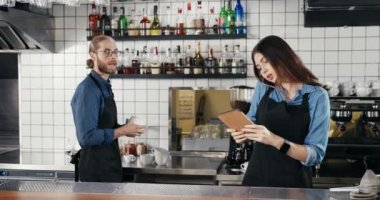 Güzel beyaz kadın barmen cep telefonuyla konuşuyor ve barda tablet cihazını tutuyor. Barikatlar arka planda çalışıyor. Garson cep telefonuyla konuşuyor ve cihazları dinliyor.