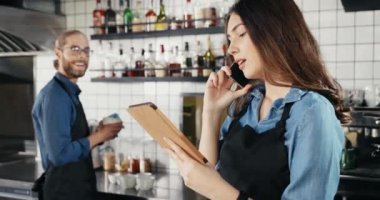 Güzel beyaz kadın barmen telefonda konuşuyor ve barda tablet cihazını tutuyor. Adam arka planda çalışıyor ve kıza gülümsüyor. Garson kız cep telefonuyla konuşuyor.