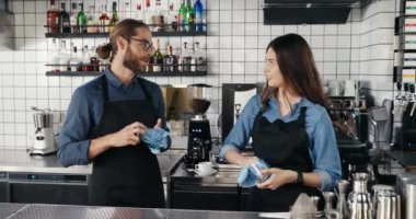 Kafede gülen, sohbet eden ve bardakları silen önlüklü, beyaz erkek ve kadın bir çift. Mutlu garson ve garson bardakları ve kurutucuyu temizliyor. Barristas neşeyle çalışıyor.