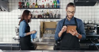 Kafede tablet kullanan gözlüklü ve önlüklü beyaz bir erkek. Bar garsonluğu yapan yakışıklı bir adam. Cihazla internette geziniyor. Garson, arka planda çalışıyor. Kadın gözlüğü..