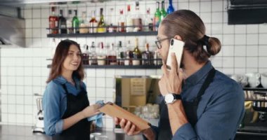 Yakışıklı beyaz bir barmen cep telefonuyla konuşuyor ve barda tablet cihazını tutuyor. Barikat elemanı cep telefonundan konuşuyor ve arka plandaki garsona dönüyor. Kadın iş arkadaşı..