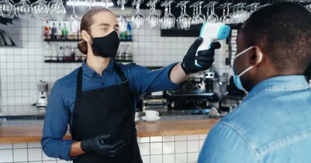 Pria kulit putih bartender tampan bertopeng dan sarung tangan mengukur suhu untuk klien Afrika-Amerika di kafe. Melepaskan pelanggan di bar. Konsep Coronavirus. dibuka kembali setelah proses karantina selesai.. — Stok Video