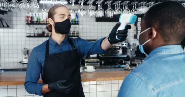 Maschio caucasico bel barista in maschera e guanti che misura la temperatura al cliente afroamericano nel caffè. Lasciar andare i clienti al bar. Concetto di Coronavirus. riaprire dopo covid quarantena blocco. — Video Stock