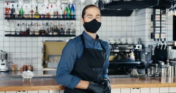 Kafenin tezgahında durup kameraya bakan maskeli ve eldivenli beyaz bir adamın portresi. Barda önlüklü yakışıklı bir erkek. Covid-19 karantina konsepti. — Stok video