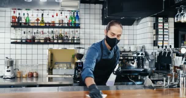 Кавказький самець бармен в масці і рукавичках витирає поверхню стійки бару дезінфектором. Чоловік чекає на прибирання стола в кафе, розбризкуючи антибактеріальну рідину. Дезінфекція громадського місця. Корона. — стокове відео