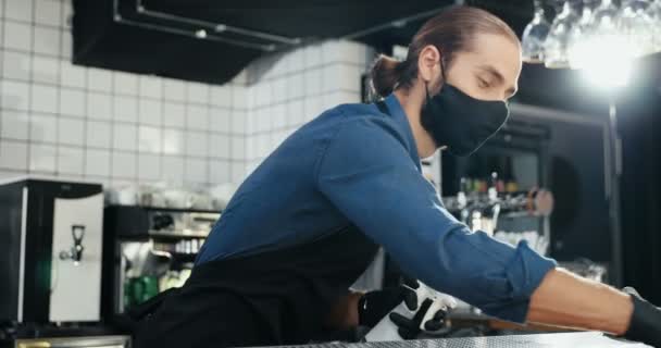 Barman maschio caucasico in maschera e guanti pulire superficie del bancone bar con disinfettore. Cameriere uomo tavolo di pulizia in caffè, spruzzando liquido antibatterico. Disinfezione del luogo pubblico. Misure transitorie. — Video Stock