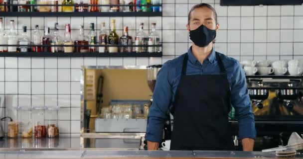 Kafe 'de maske, eldiven ve önlük takmış beyaz, yakışıklı bir adamın portresi. Erkek barmen barda tezgaha yaslanırken dönüp düz bakıyor. Coronavirüs salgını konsepti yeniden aç. — Stok video