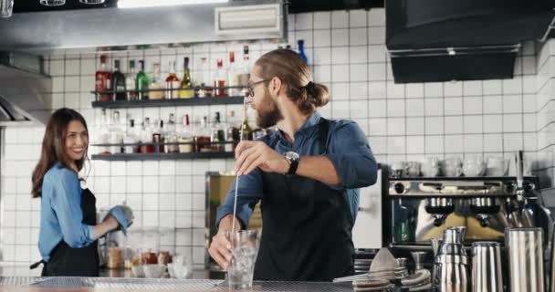 Caucasiano jovem bonito e alegre barman preparando bebida alcoólica em rochas e sorrindo para colega mulher. Garçom e garçonete falando. Guy barrista fazendo coquetel com gelo no bar. — Vídeo de Stock