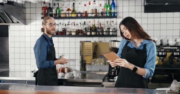 Prachtige jonge vrouwelijke blanke barman die praat op een mobiele telefoon en een tablet vasthoudt aan de bar. Man Barrista werkt op de achtergrond. Serveerster spreekt op mobiele telefoon en tikt op gadget — Stockvideo