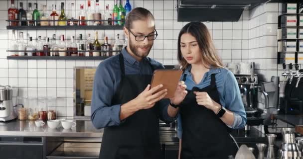 Barristas caucásicos jóvenes, alegres, hombres y mujeres, de pie en la cocina del bar y utilizando un dispositivo de tableta. Camarero y camarera viendo vídeo y tocando en la computadora gadget en la cafetería. Cantineros. — Vídeos de Stock