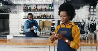 Güzel Afro-Amerikalı garson barda cep telefonunu dinleyip kaydırıyor. Kahveci kadın akıllı telefondan mesaj atıyor. Garson arka planda bardakları siliyor. Kafede temizlik bardakları.