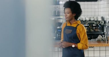Güzel Afrikalı Amerikalı garson kameraya doğru dönüyor ve tezgahta mutlu bir şekilde gülümsüyor. Kadın kahveci kameraya poz veriyor. Garson arka planda bardakları siliyor. Kafede temizlik bardakları.