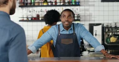 Afro-Amerikalı garson kafede beyaz bir adamla gülüyor ve konuşuyor. Erkek barikatı müşteriyle güzel bir konuşma yapıyor. Erkek barmen erkek müşteriyle sohbet ediyor. Karışık ırklar. Çoklu Etnik