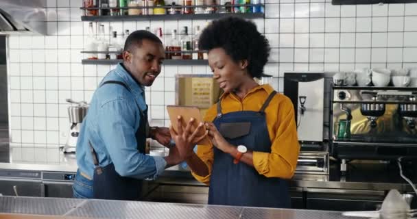 Önlüklü güzel Afro-Amerikalı bayan garson tezgahta duruyor, tablet cihazı kullanıyor ve erkek garsonla konuşuyor. Barmen 'e barmen gösterirken kadın barfiks tıkırdıyor.. — Stok video