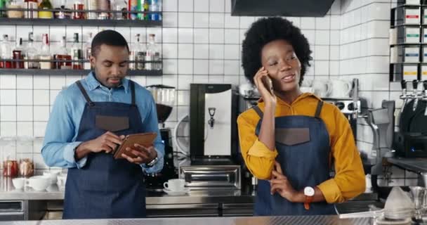 예쁘게 생긴 아프리카계 미국인 여성 웨이트리스가 카운터에서 카페에 서서 핸드폰으로 얘기하고 있습니다. 남자 바리스타가 태블릿으로 문자를 보내고 스크롤을 하고 있어요. 휴대폰으로 통화하는 모습 — 비디오