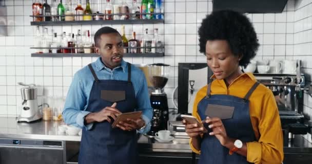 Hermosa camarera afroamericana en delantal de pie en la cafetería en el mostrador y mensaje de texto en el teléfono inteligente. Hombre barrista tocando y desplazándose en el dispositivo de la tableta. Sms al teléfono. Chatear en línea. — Vídeos de Stock