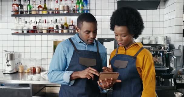 Afro-Americanos alegres abogados masculinos y femeninos de pie en la cocina del bar y el uso de tableta dispositivo. Camarero y camarera viendo vídeo y tocando en la computadora gadget en la cafetería. Cantineros. — Vídeos de Stock