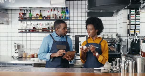 Afrikai amerikai jóképű fiatal férfi csapos kötényben tartó tabletta eszköz és női barrista írási lista megrendelések vagy vásárlás a bárban konyha. Pincér és pincérnő készítik a kávézó étlapját. — Stock videók