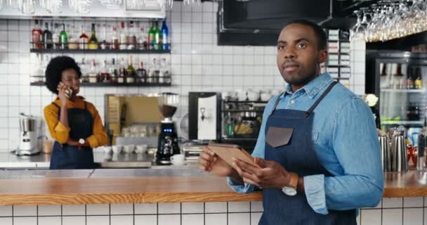 Homens barmen afro-americanos usando dispositivo tablet no café. Bonito garçom homem tocando e rolando on-line no gadget. Garçonete falando no celular no fundo. Mulher falando no celular. — Vídeo de Stock