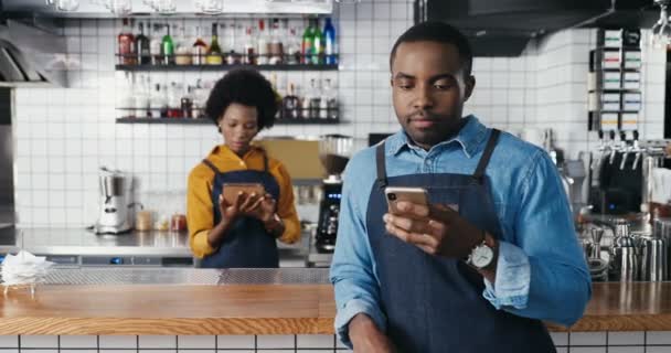 Barmen afro-americano masculino usando smartphone no café e mensagem de texto. Bonito garçom batendo e rolando on-line no telefone. Garçonete com dispositivo tablet em segundo plano. Mulher segurando gadget. — Vídeo de Stock