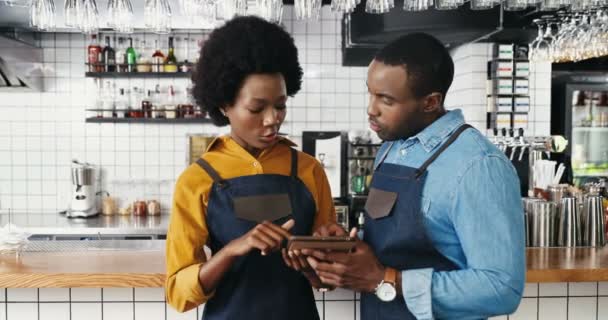 Afro-Amerikaanse vrolijke mannelijke en vrouwelijke barrista 's die in de barkeuken staan en een tablet gebruiken. Ober en serveerster kijken naar video en tikken op gadget computer in café. Bartenders. — Stockvideo