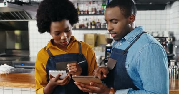 アフリカ系アメリカ人のハンサムな若い男性バーテンダーエプロン保持タブレットデバイスとバーキッチンでの注文やショッピングの女性のバリスタの書き込みリストです。ウエイターとウエイトレスがカフェのメニューをメモ — ストック動画